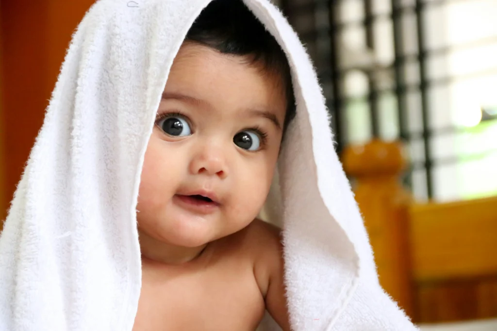 Cute baby with big eyes peeking out from a white towel, looking curious and playful.