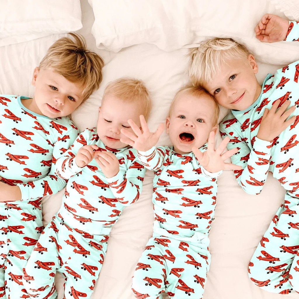 four cute toddlers in matching airplane pajamas, lying on a bed and smiling at the camera