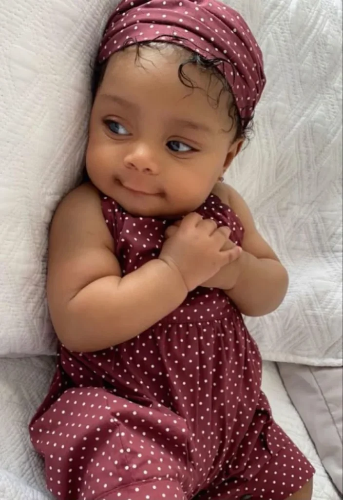 cute baby with curly hair wearing a maroon polka dot outfit, smiling and resting on a white blanket