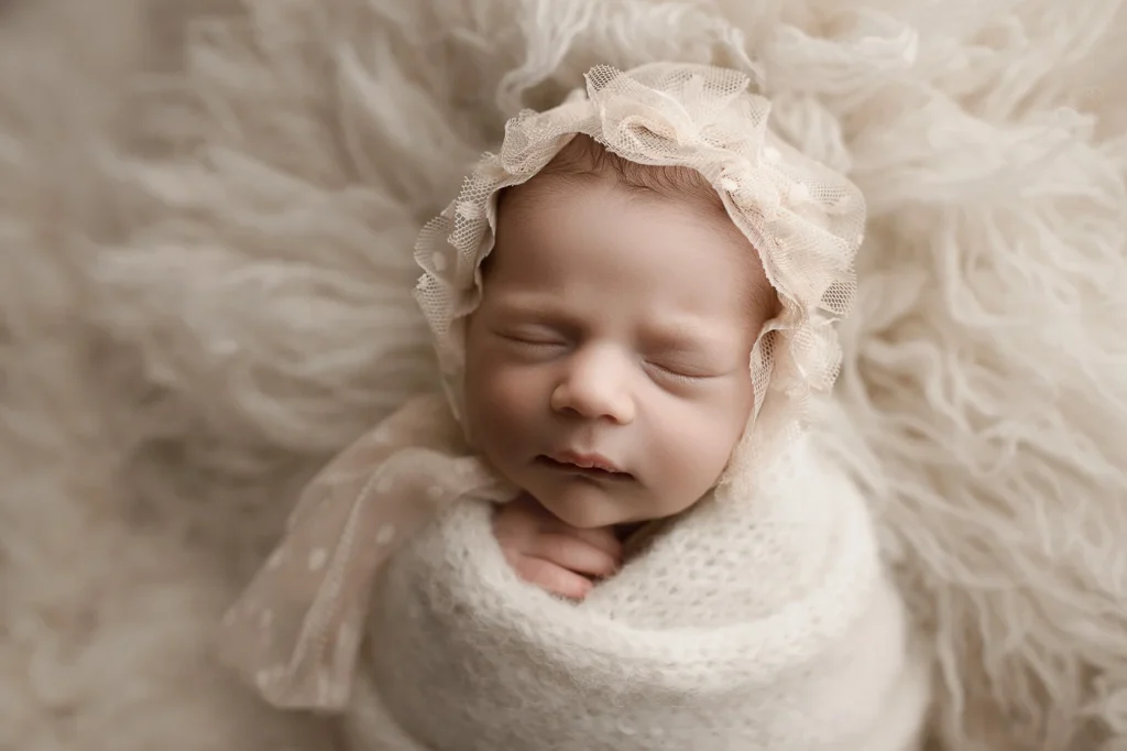 Cute baby swaddled in soft fabric, eyes closed, resting on a fluffy white surface.