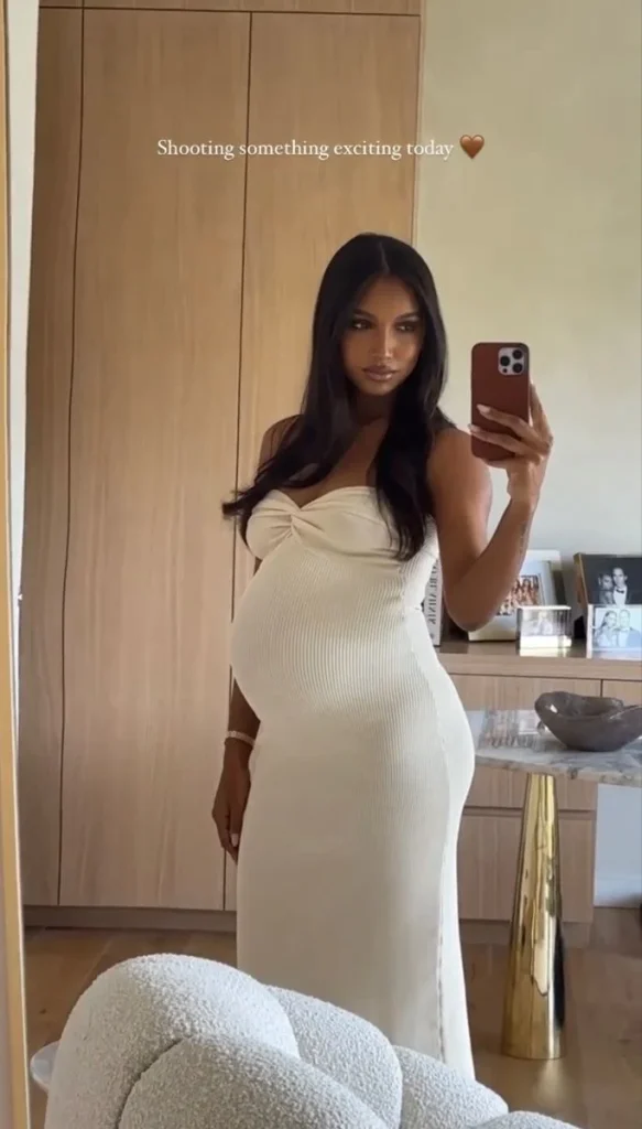 Pregnant woman in a fitted white dress taking a selfie in a stylish interior.