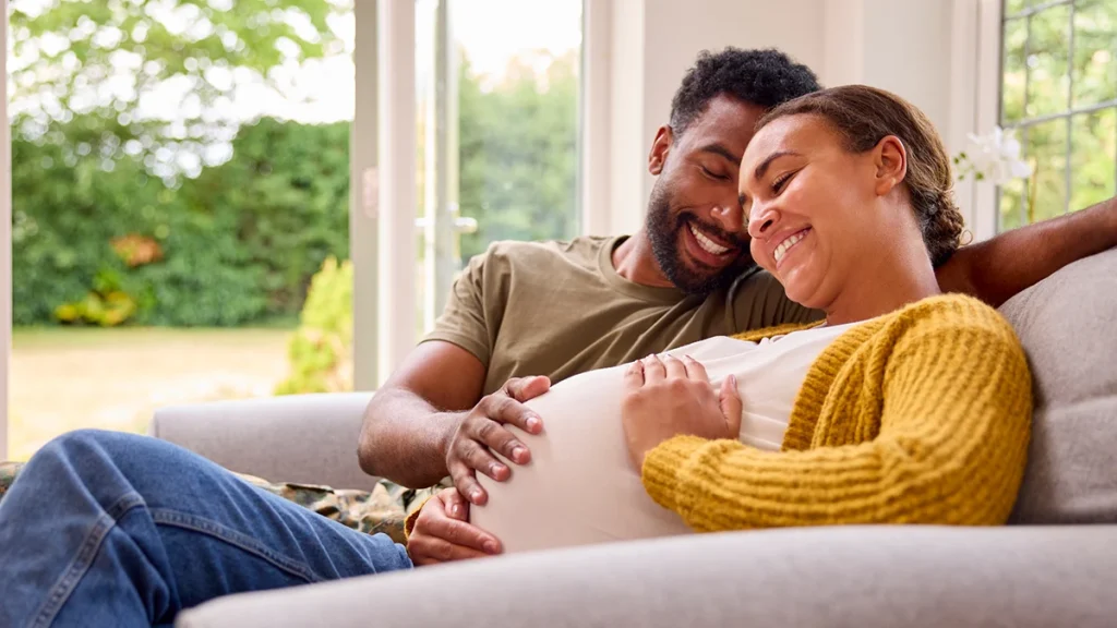 pregnant couple sharing a joyful moment together on a couch, with the woman resting her hand on her belly.